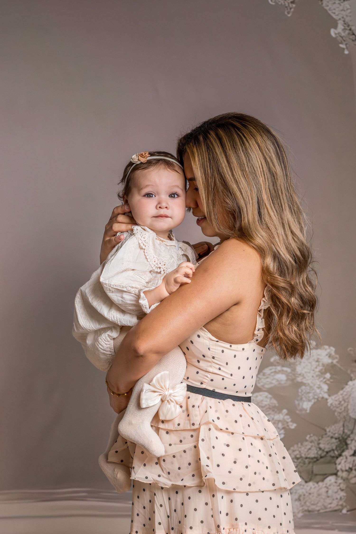 A mother in a polka dot dress tenderly kisses her baby girl in a white dress and bow headband in a studio.