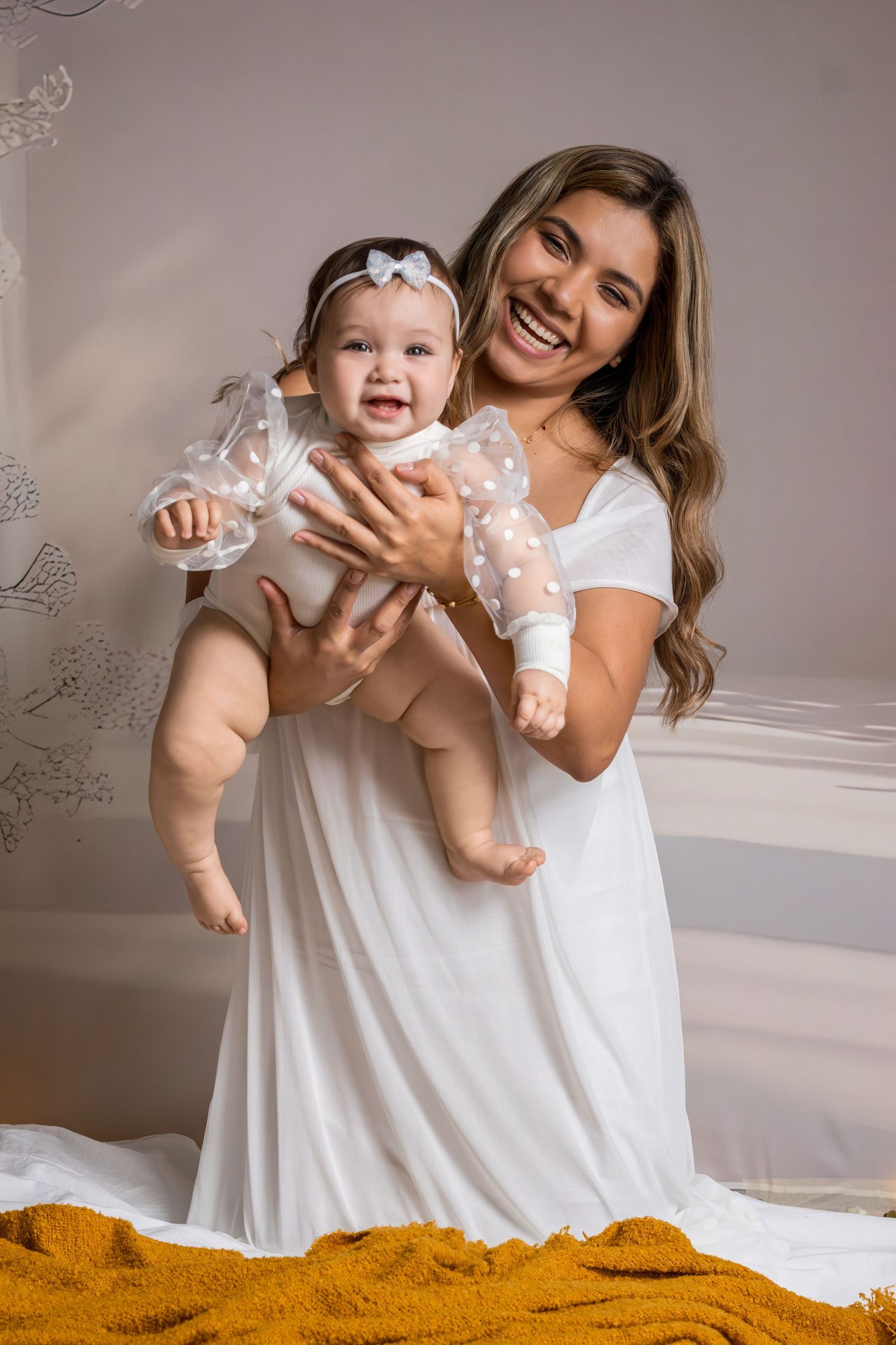 A joyful mother in a white dress holds her smiling baby girl dressed in a white polka dot outfit.