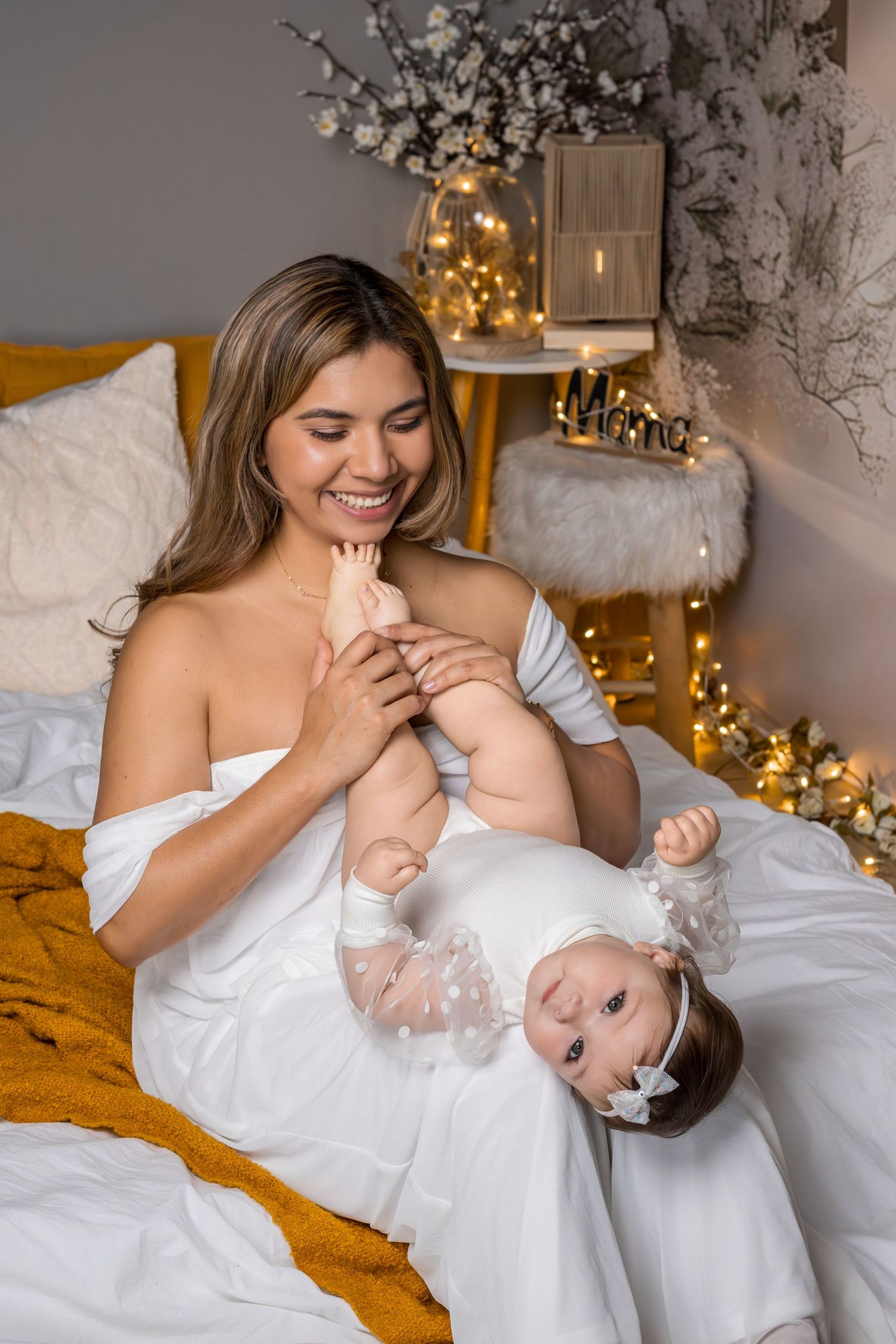 Smiling mother in white dress holding her baby girl in a festive bedroom with warm golden lights.