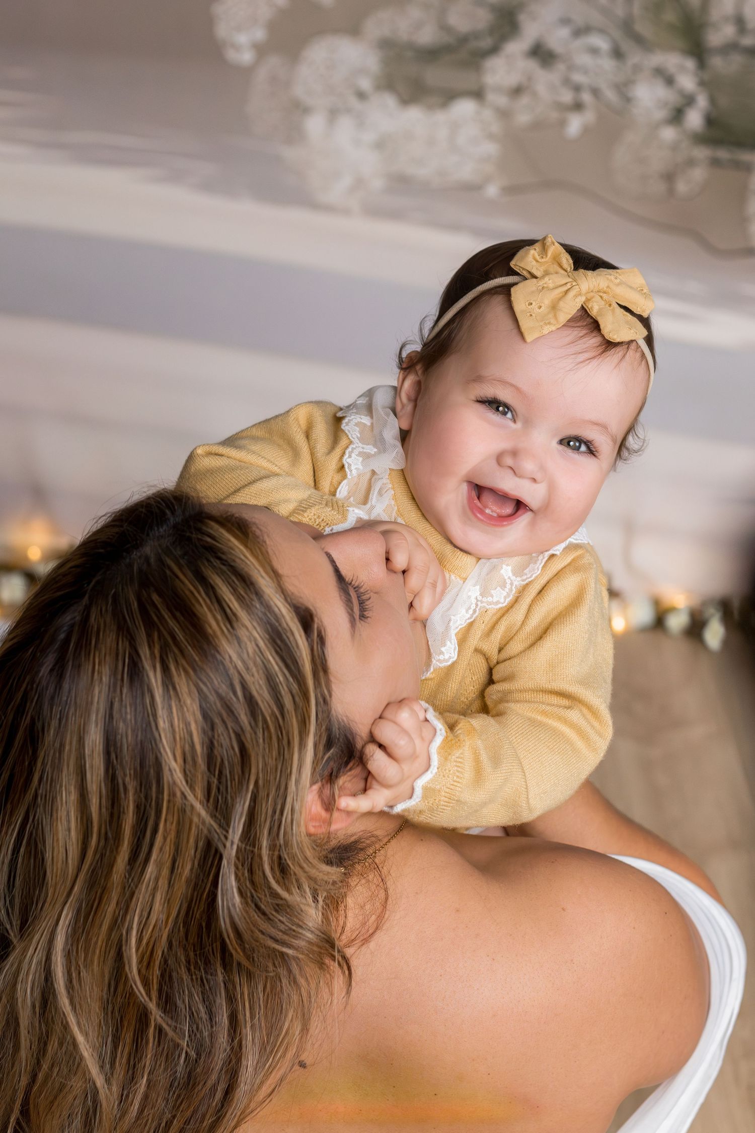 Mother kissing her smiling baby girl in a yellow bow headband and golden outfit in a warmly decorated room.