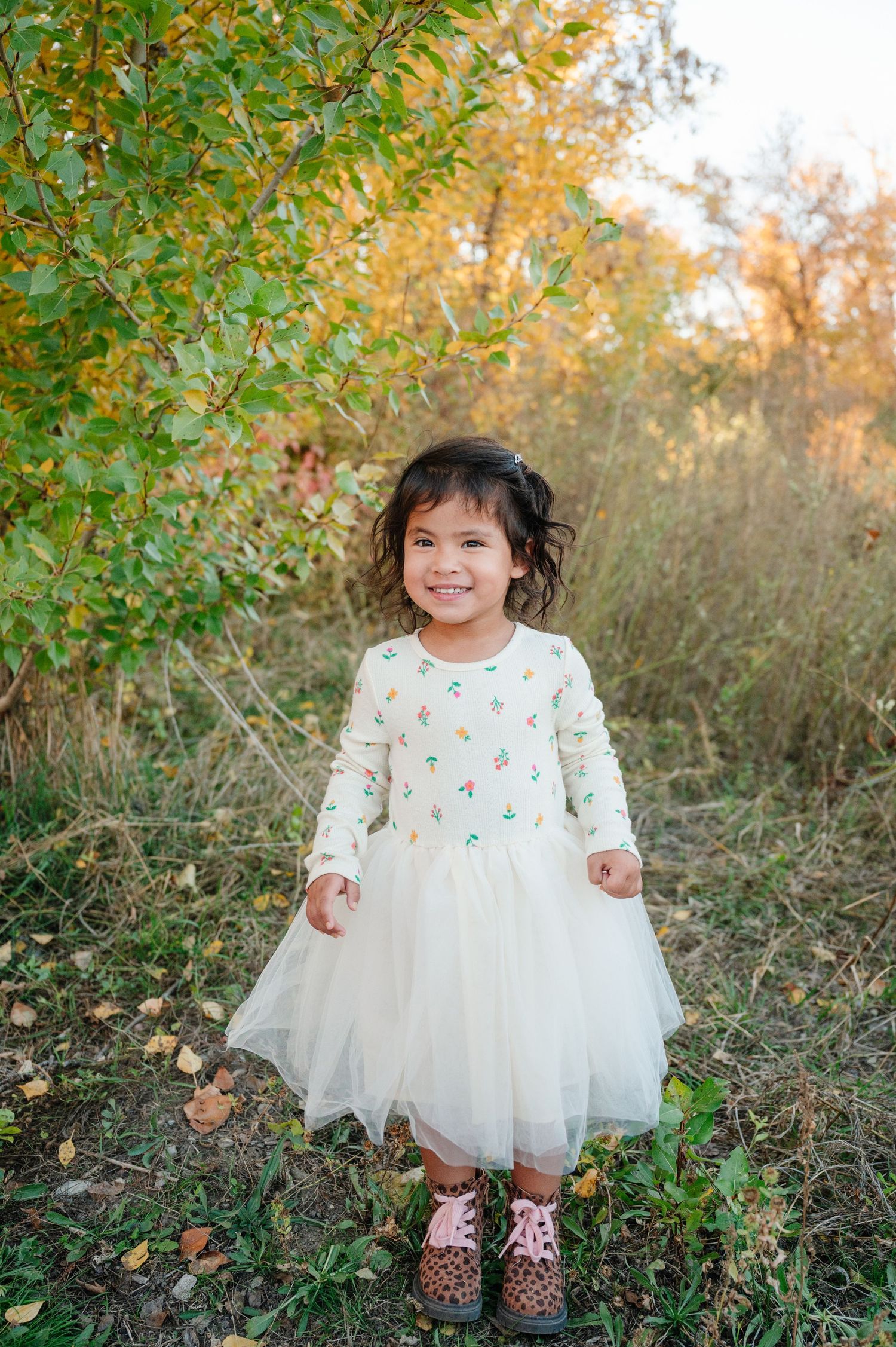 Young child in white tulle dress and sparkly shoes stands among autumn foliage.