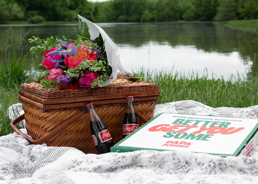 Picnic setup with wicker basket, flowers, and blanket by a serene lake.