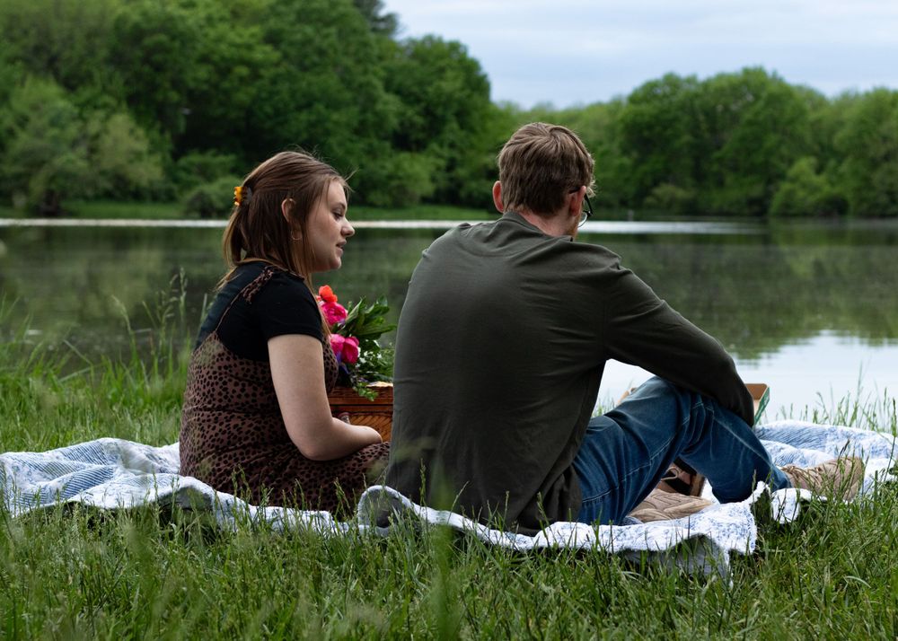 Friends lounging on blanket beside lake with picnic setup.