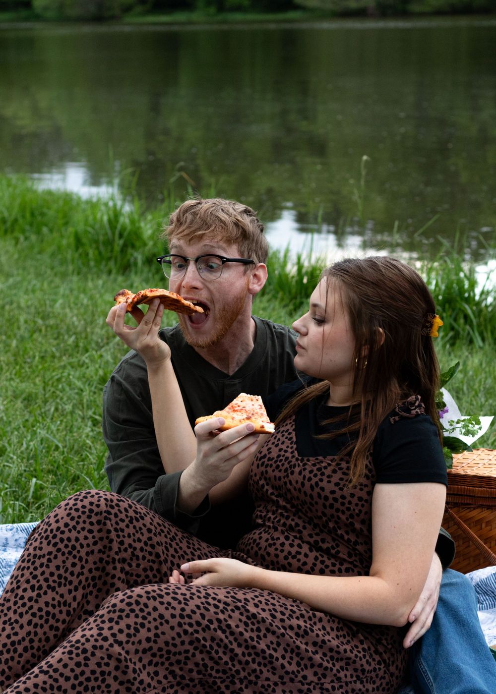 Two people laughing and eating pizza together during a lakeside picnic.