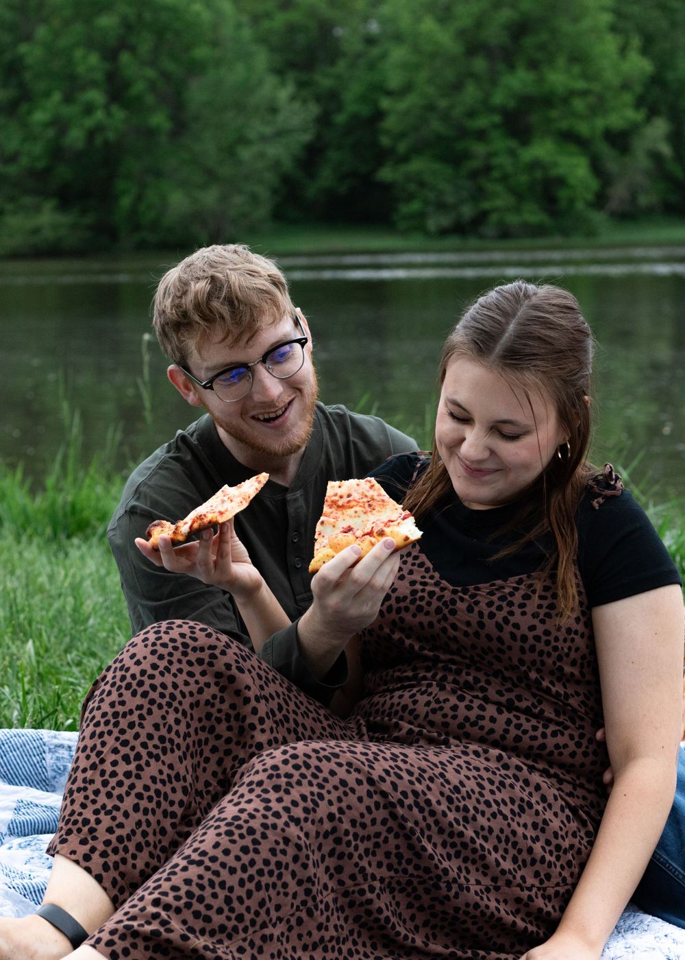 Two people sharing pizza and laughing during a romantic picnic by the water.