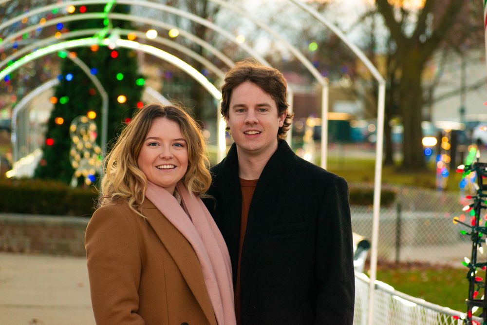 A couple poses under an illuminated archway decorated with holiday lights.