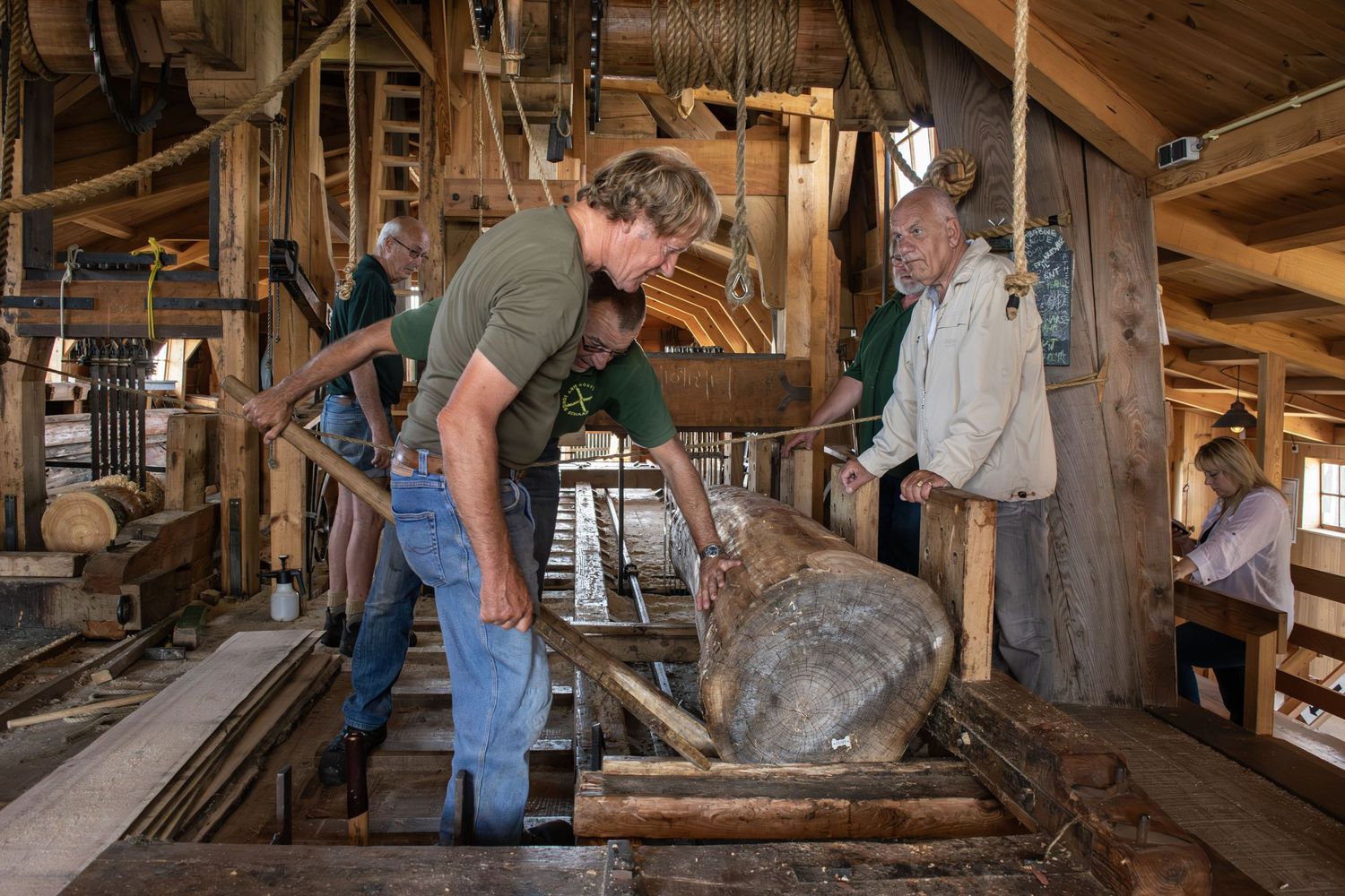 Historic mill workers examining large wooden equipment in rustic interior.