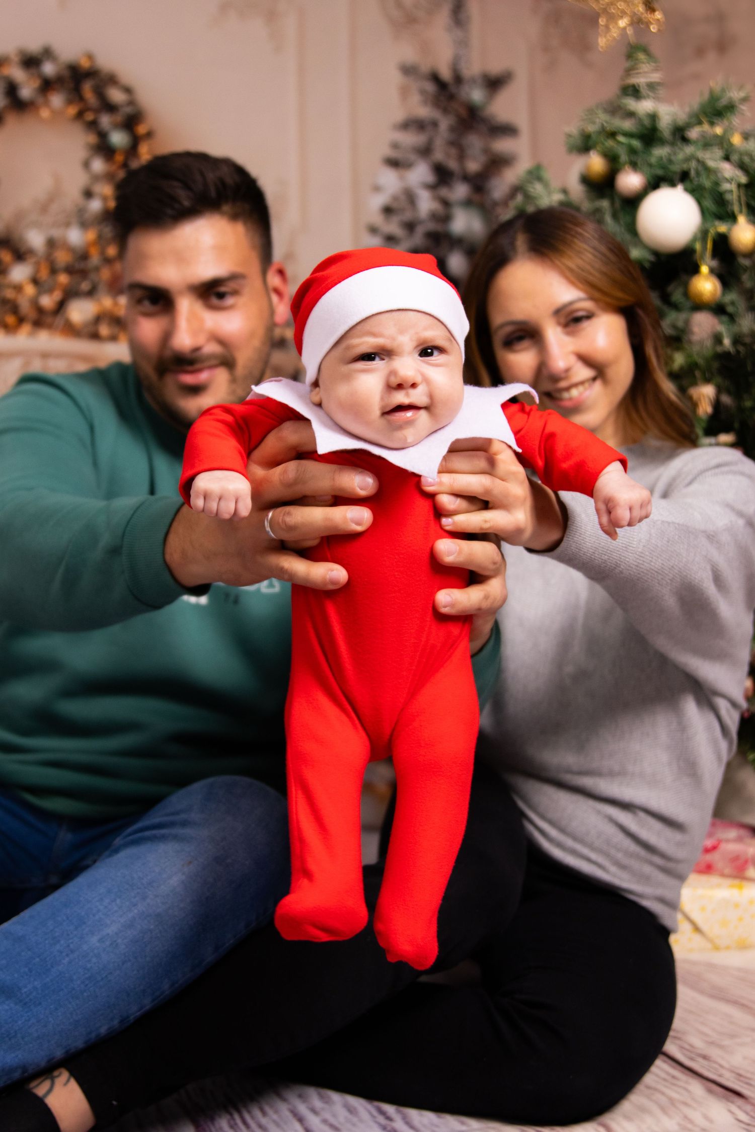 Family playing with baby in red Christmas outfit on couch with decorated tree in background.