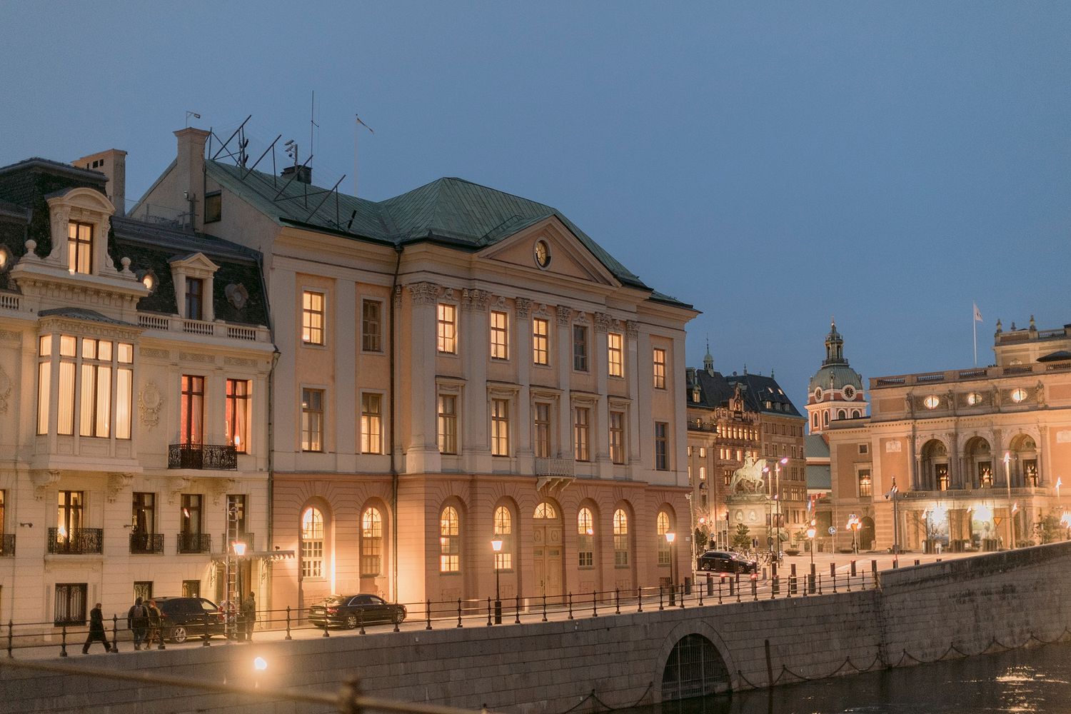 Elegant historic building illuminated at dusk along a waterfront promenade.