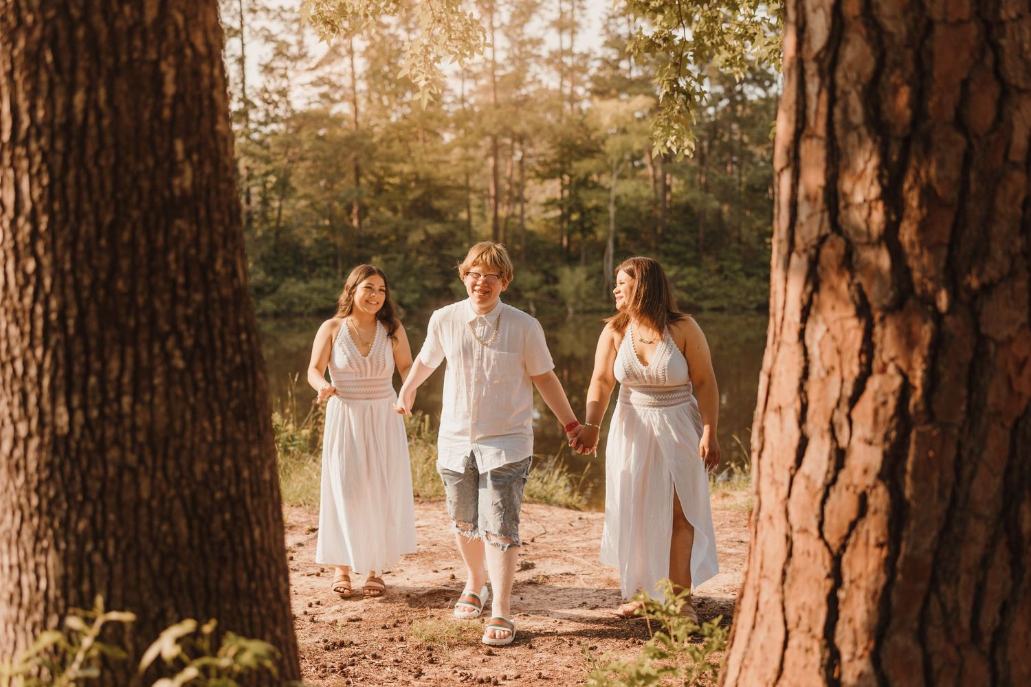 Three people in flowy white outfits walk together through a sun-dappled forest path.