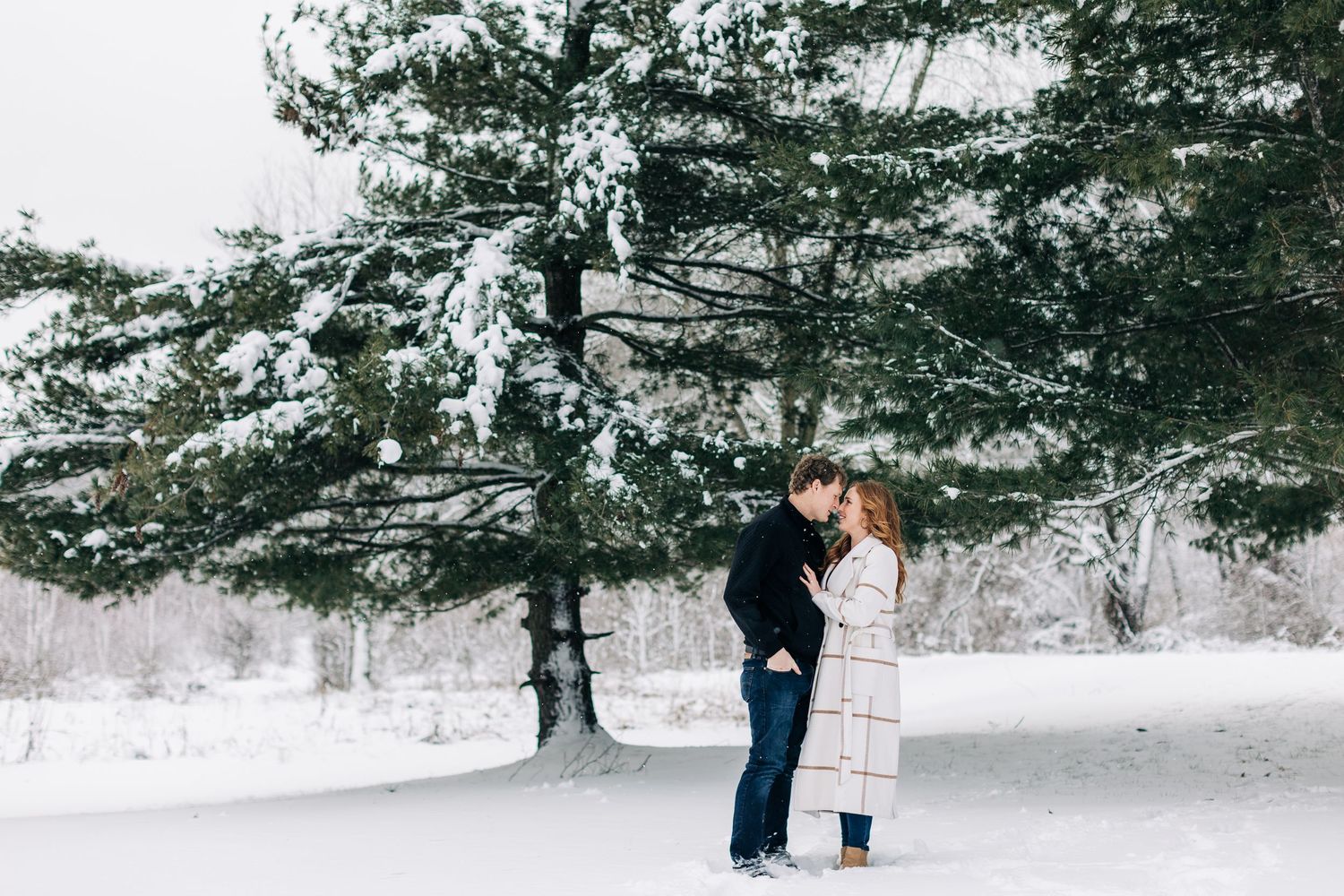 Romantic couple stands close together in snowy winter scene with evergreen trees.
