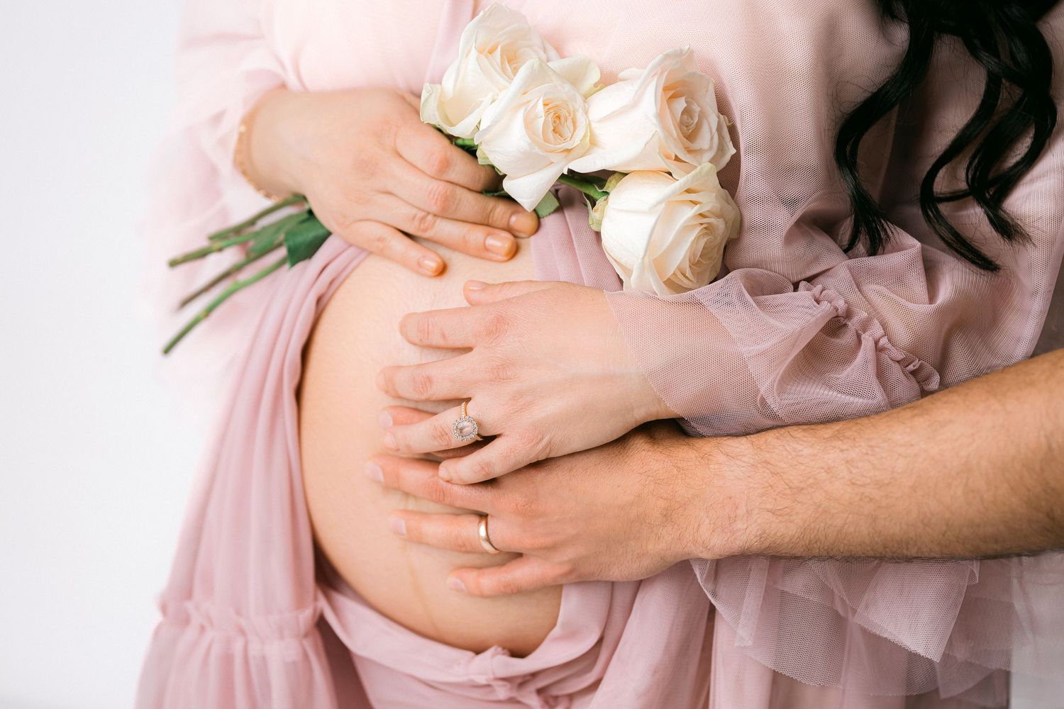 An elegant maternity portrait with white roses and a flowing pink lace dress against a neutral background.