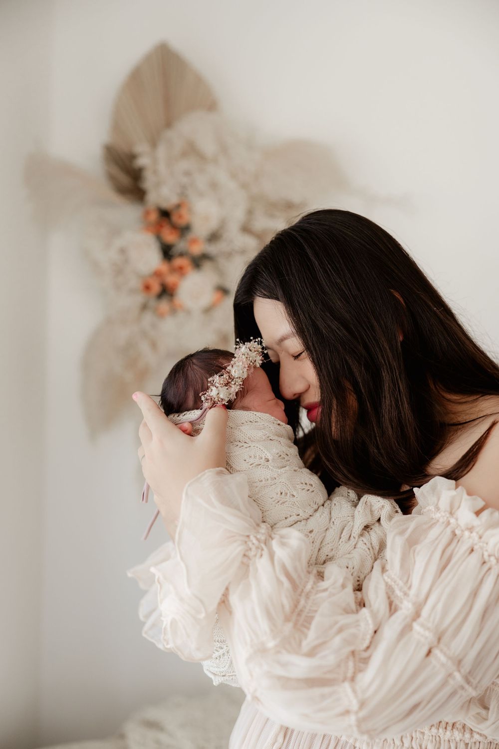 A mother in a white lace dress tenderly holds her newborn baby near a boho floral wall decoration.