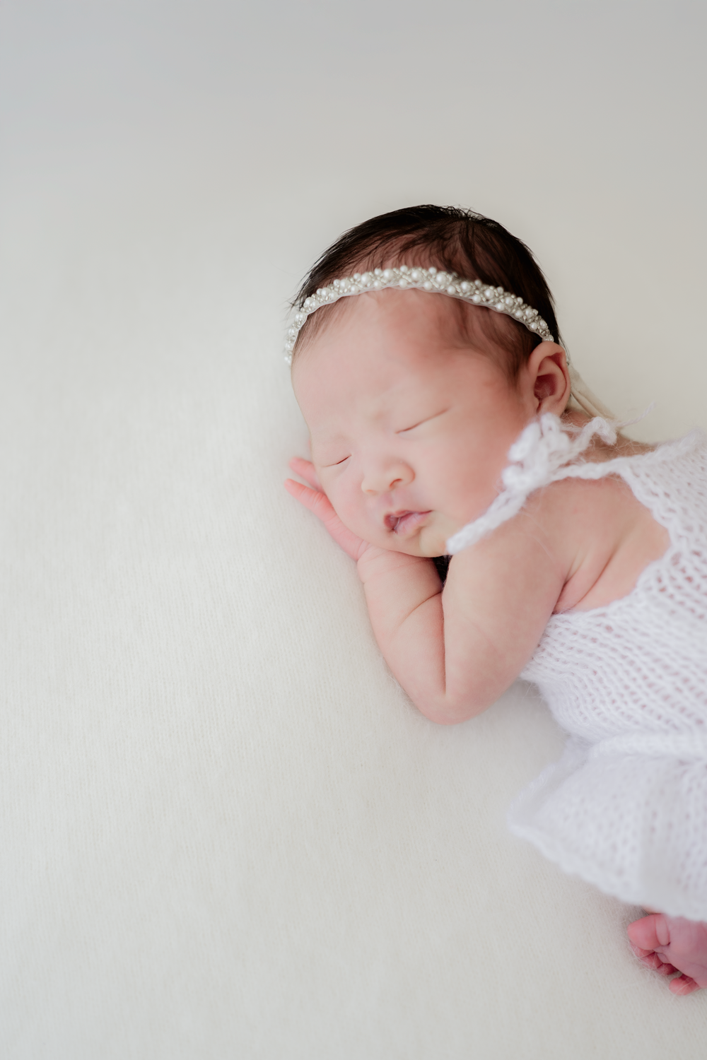 Sleeping newborn baby girl in white knit outfit and pearl headband on soft white background.