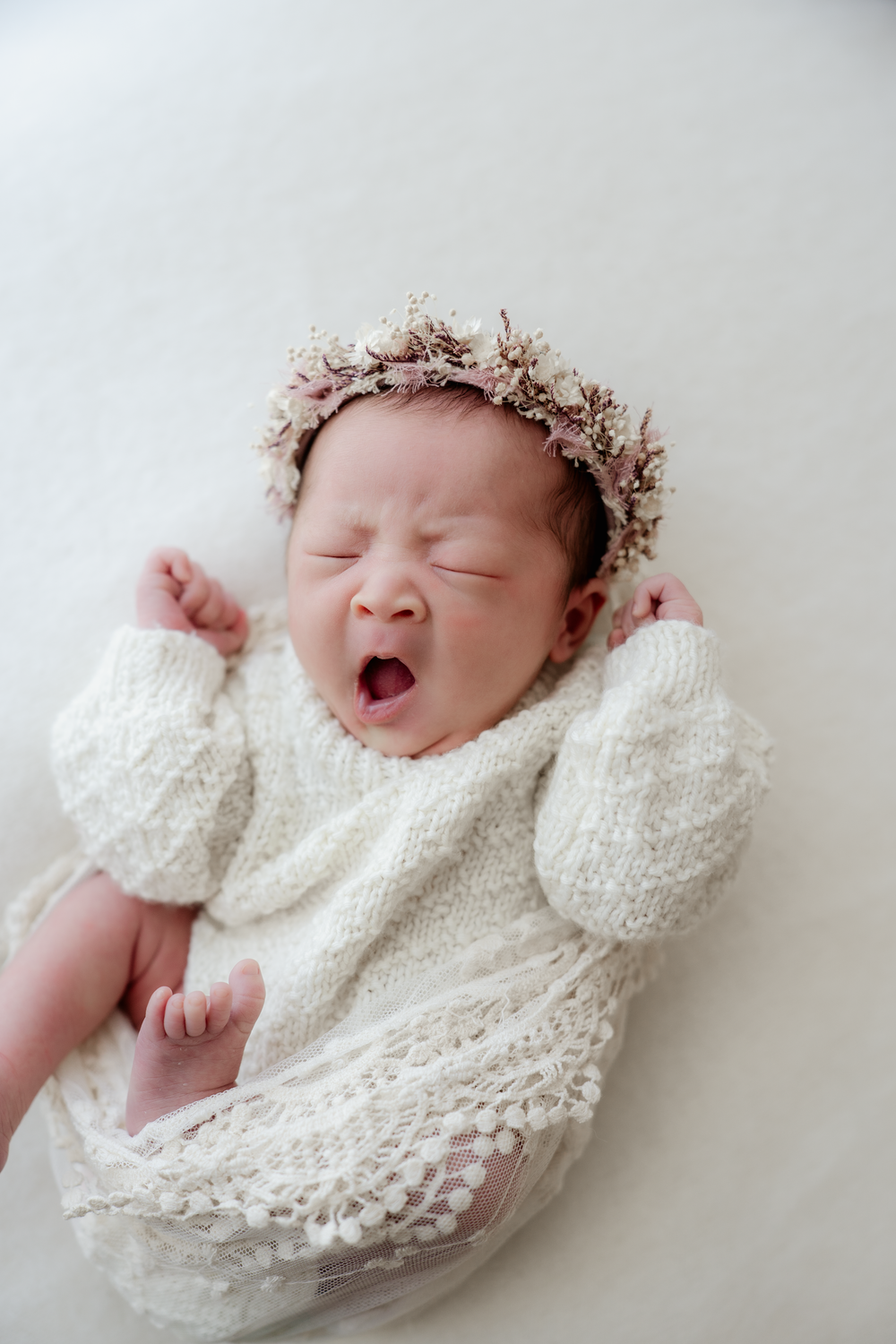 Newborn baby yawning in white knit outfit and floral crown, lying on soft white blanket.