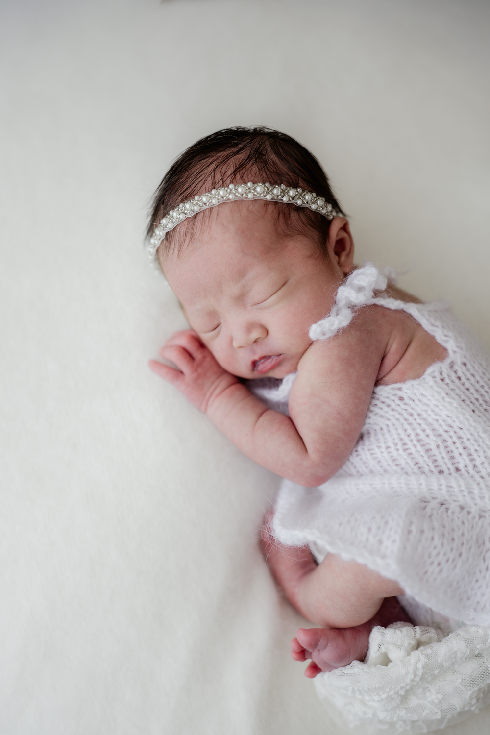 Sleeping newborn baby girl in white knit outfit and rhinestone headband lying on soft white blanket.
