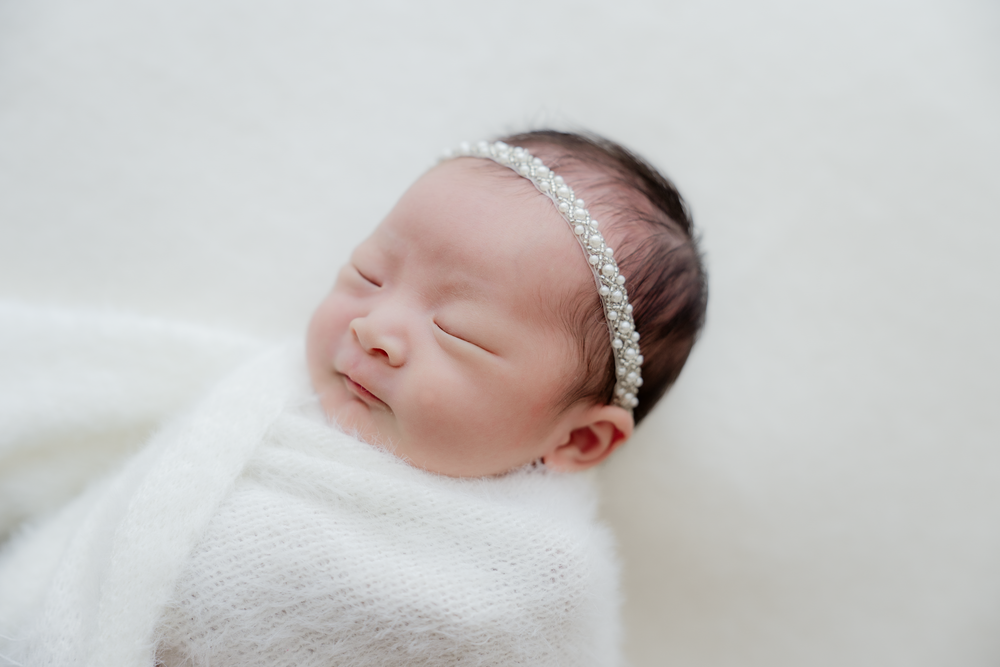 Peaceful newborn baby sleeping wrapped in white swaddle with pearl rhinestone headband on soft white background.