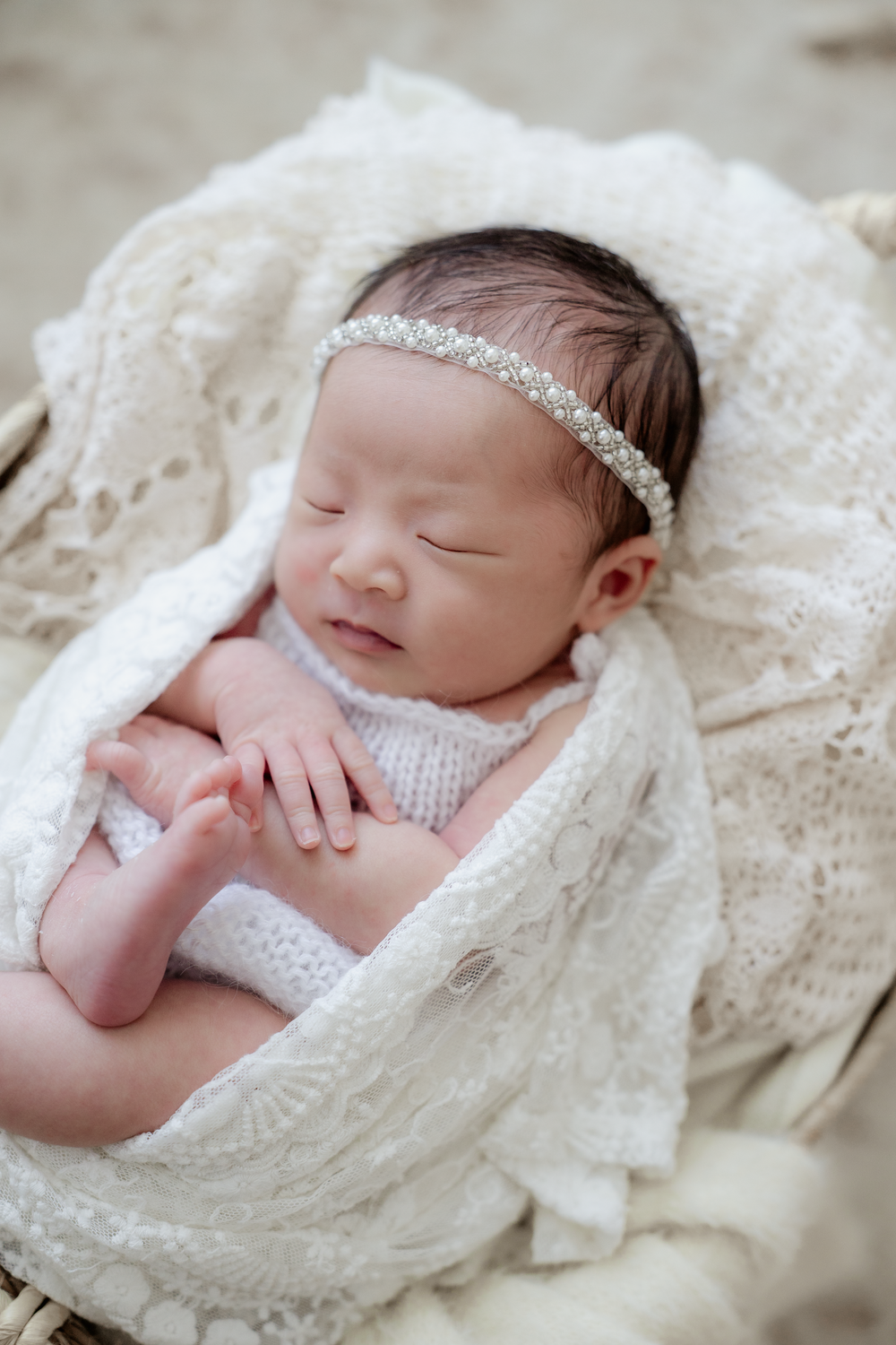 Sleeping newborn baby girl wrapped in white lace blanket wearing a pearl headband in a wicker basket.