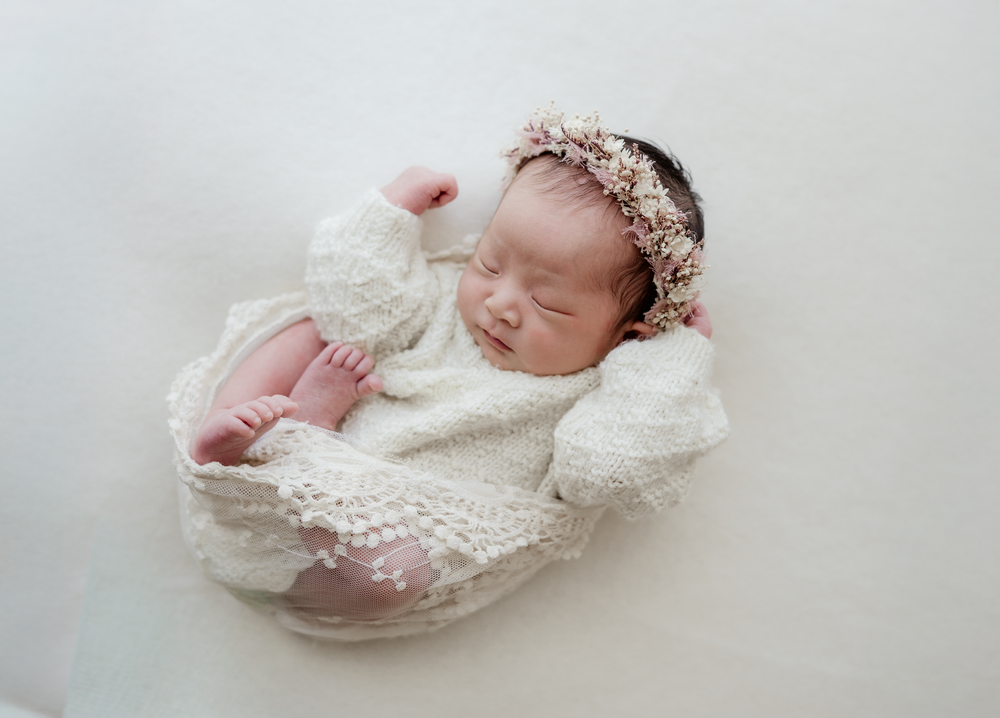 Newborn baby girl sleeping peacefully wrapped in white lace, wearing a delicate floral headband on a white background.