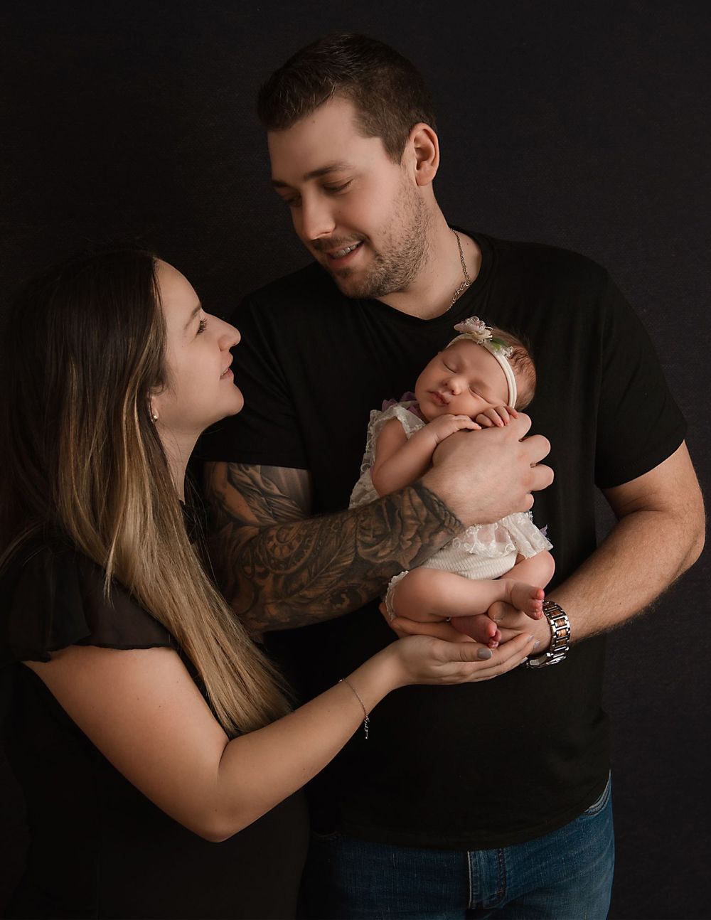 Parents share an intimate moment while holding their newborn baby in a dark studio setting.