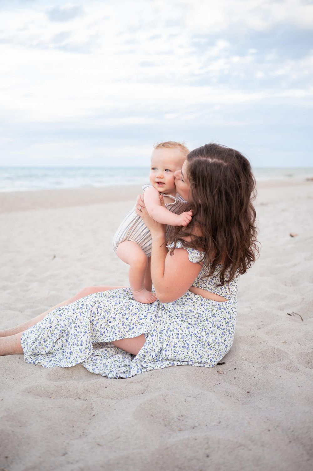 Mother kissing her baby in her lap sitting on the beach with blue sky Mommy and Me Photoshoot Melbourne Cocoa Beach Brevard County Florida