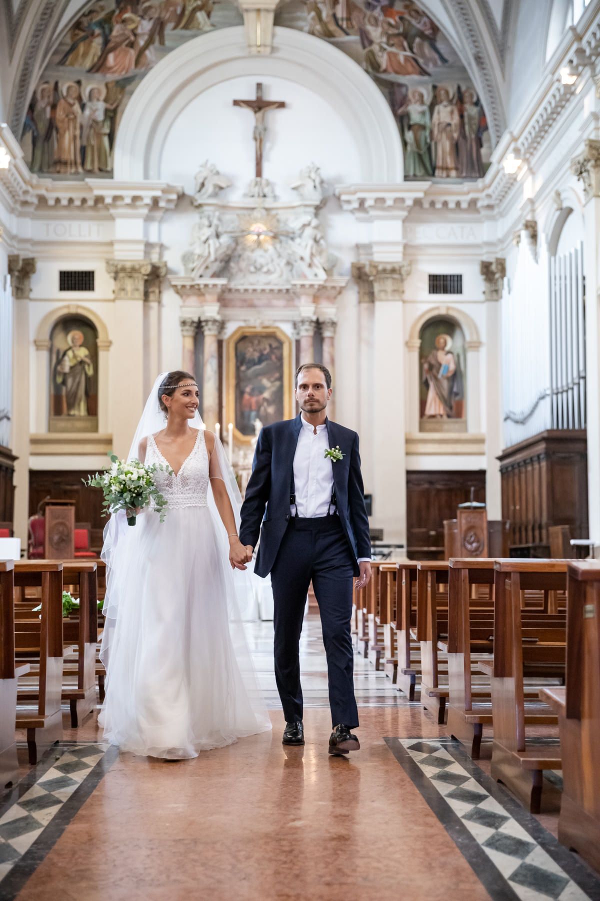 A wedding ceremony procession walking down the aisle of an ornate Catholic church with white marble columns and frescoes.