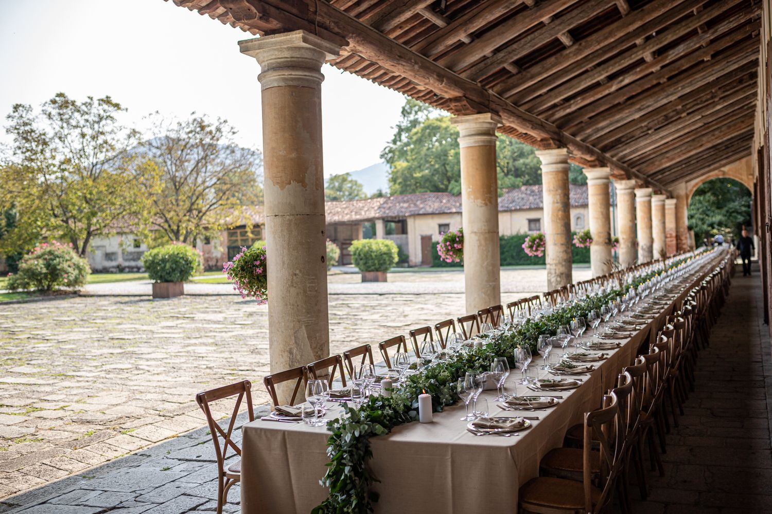 Long banquet tables set under a covered stone colonnade at an outdoor wedding venue.
