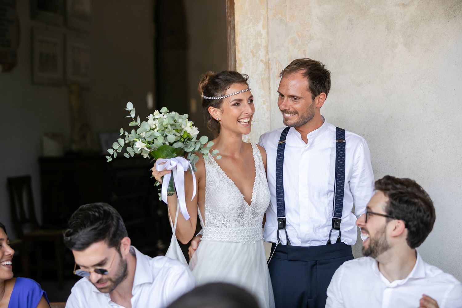 Wedding party dressed in white and suspenders posing together against a light wall.