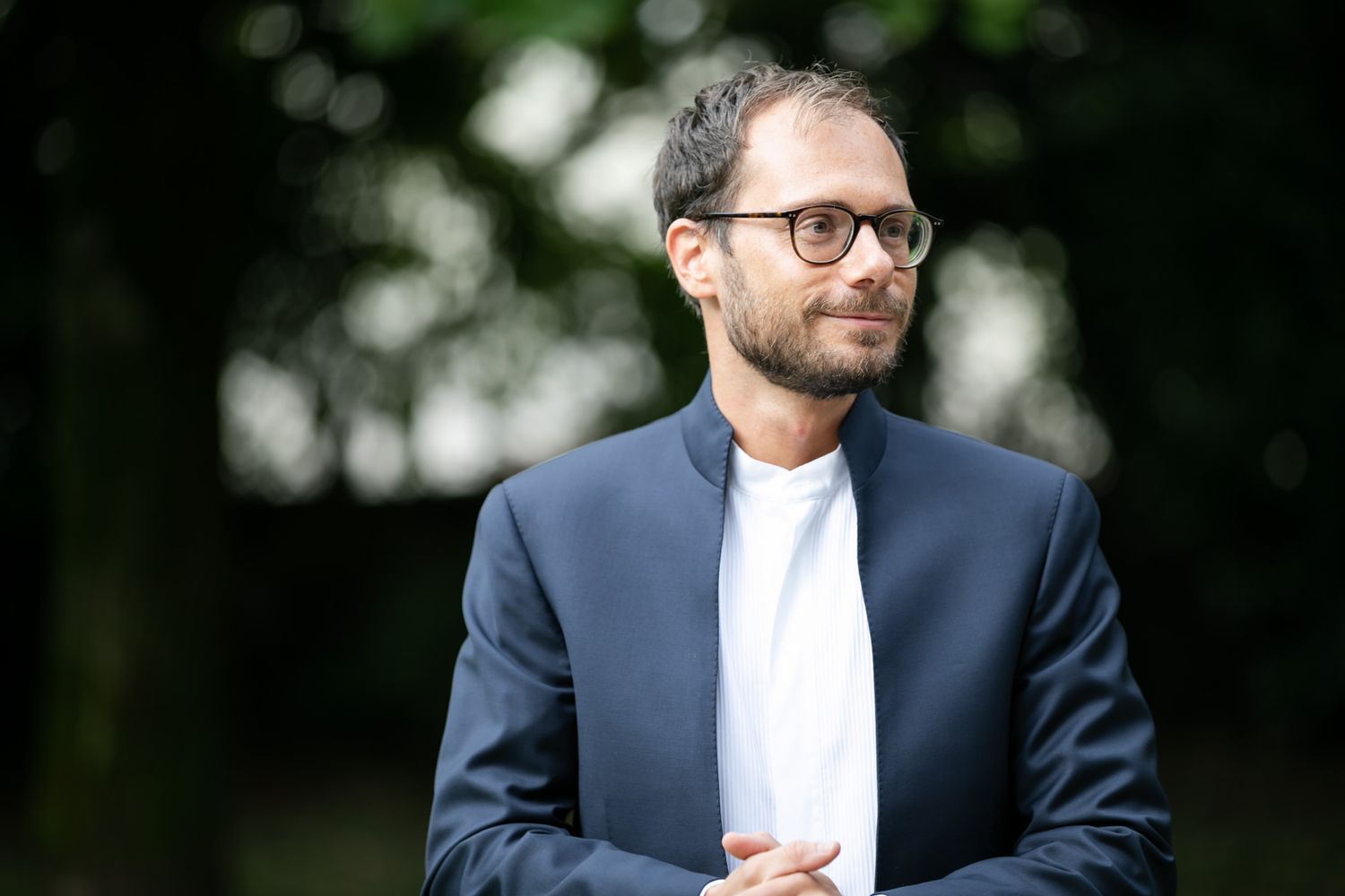 Professional portraits in a navy suit and white shirt against a blurred outdoor background.
