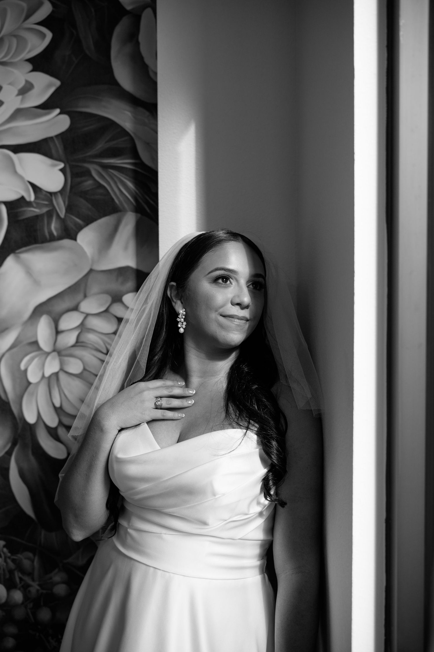 A bride in an elegant white wedding dress stands in soft window light against a floral wallpaper background.