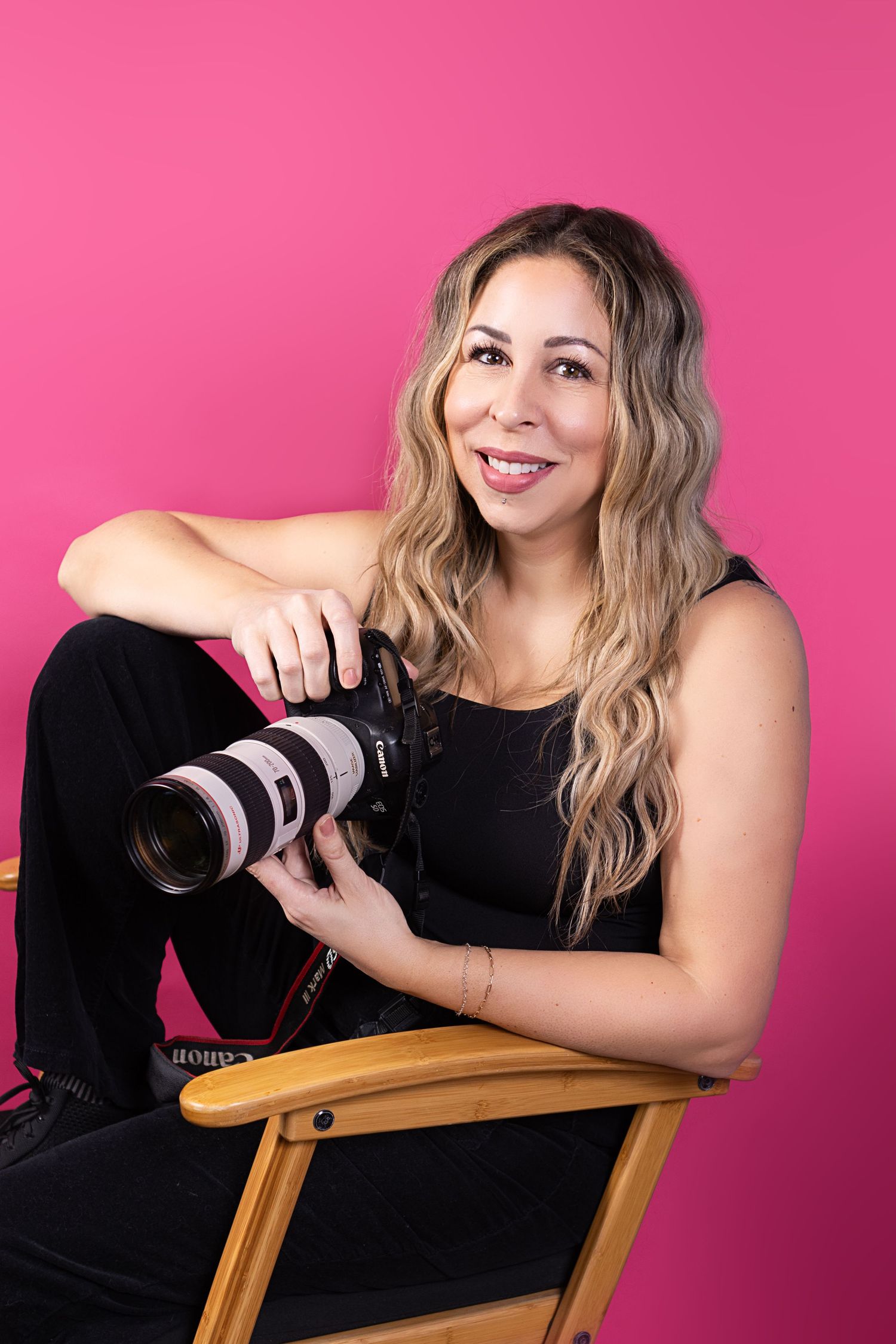 Photographer posing with a camera while sitting on a wooden chair against a bright pink background.