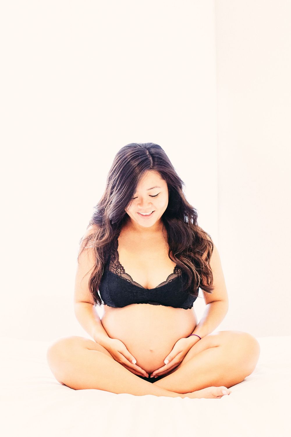 Expecting mother in black lingerie sits cross-legged making a heart shape with hands over her belly.