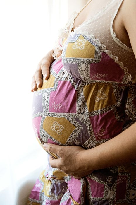 A pregnant belly shown in a patchwork dress with lace trim details against a bright window.