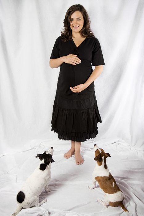 Expectant mother in black dress poses with two spotted dogs on white background.