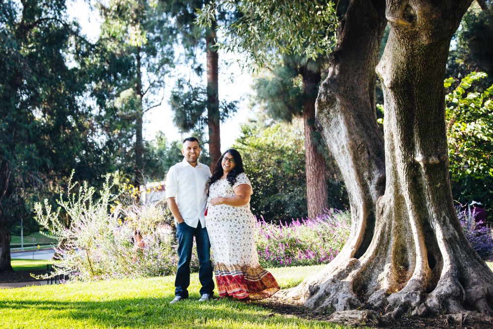A couple stands together in a sunlit garden next to a large tree trunk during a maternity photoshoot.