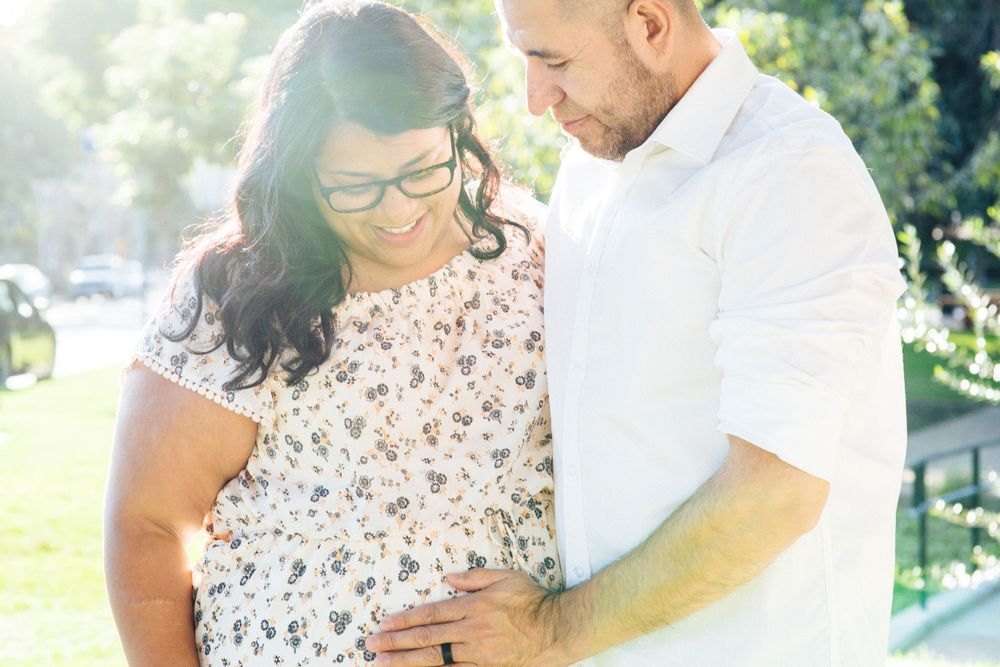 An expectant couple share an intimate moment outdoors in soft sunlight.