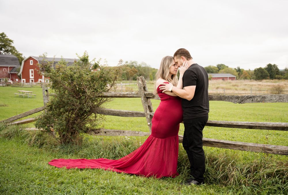 A romantic maternity photoshoot in a rustic farm setting with a flowing red dress against wooden fencing.