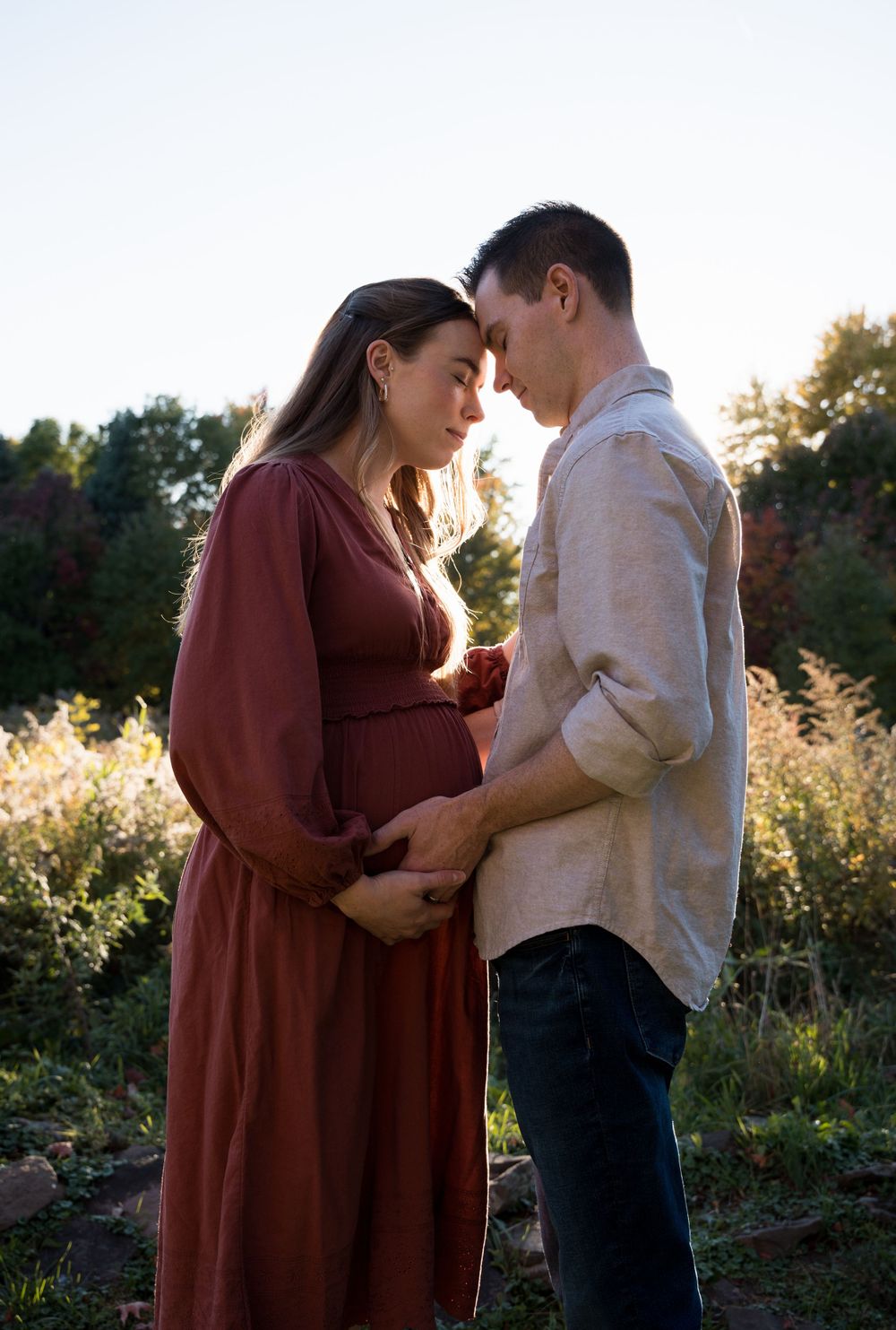 Intimate maternity portrait of couple in burgundy dress and white shirt against natural outdoor setting.