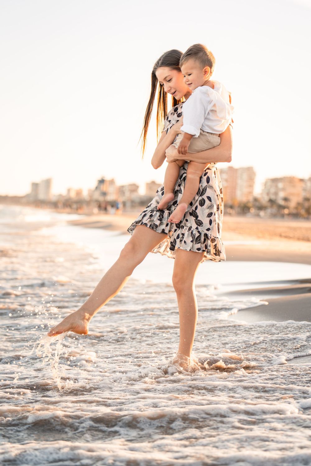 mother holding young son and splashing playfully in the water on the beach at sunset and wearing dress for family photos