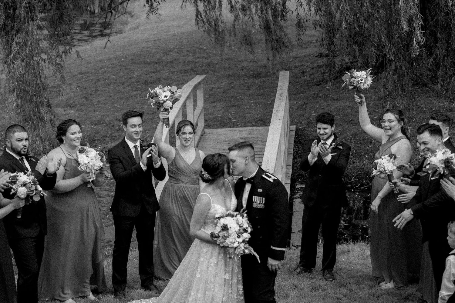 Wedding celebration scene outdoors with guests gathered under willow trees.