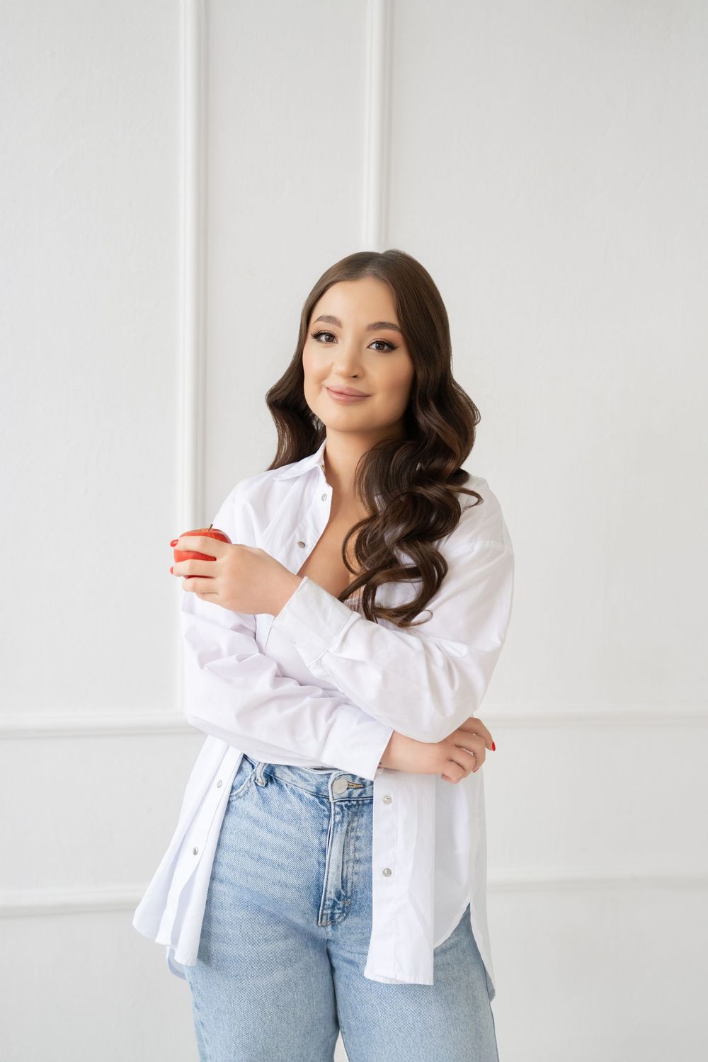 A person wearing a casual outfit with a white button-down shirt and light blue jeans poses against a white wall.