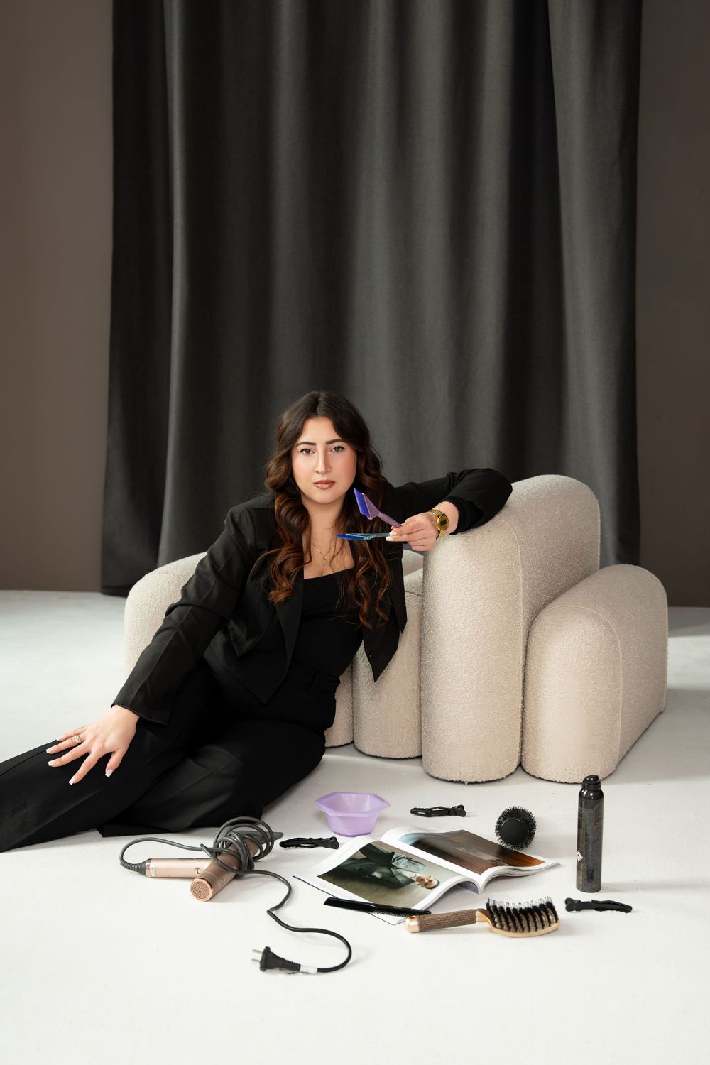 Woman in black suit reclines on beige couch with beauty tools scattered around.