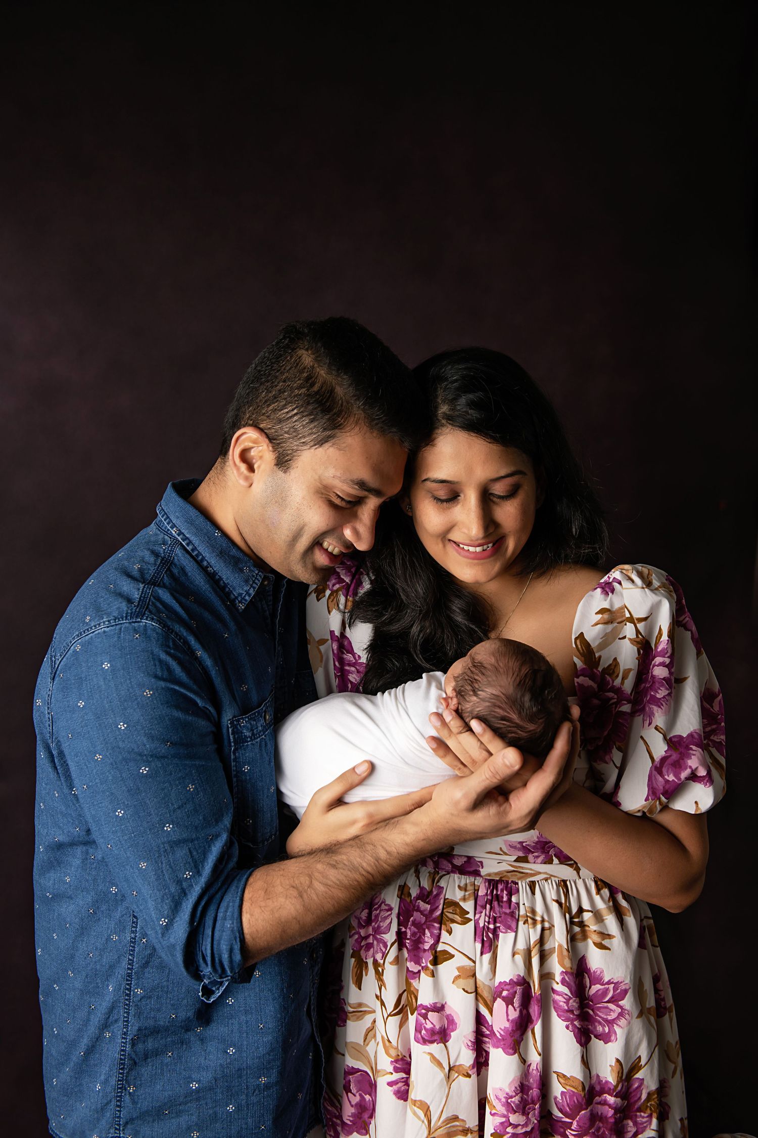 Parents in casual attire gently hold their newborn baby against dark studio backdrop.