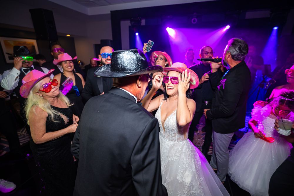 Wedding reception dance floor filled with guests wearing festive hats under purple lighting.