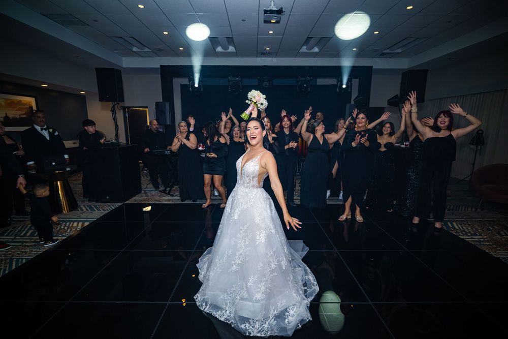 Bride in lace wedding dress poses on dark dance floor while guests cheer in background.