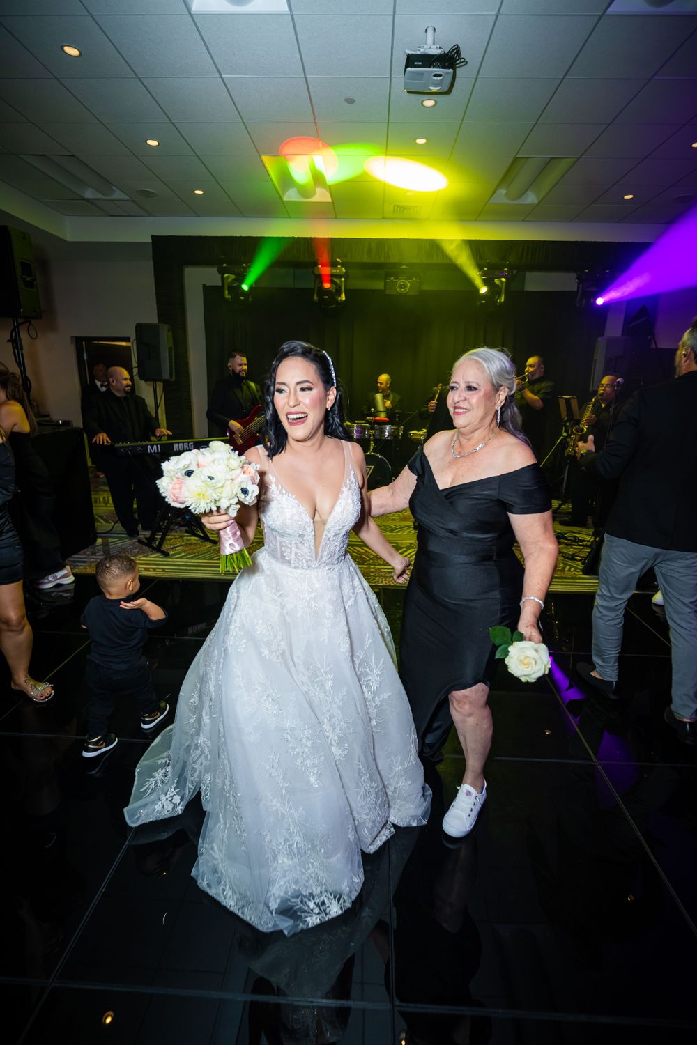 Bride in white gown and companion walk across dance floor under colorful party lights.