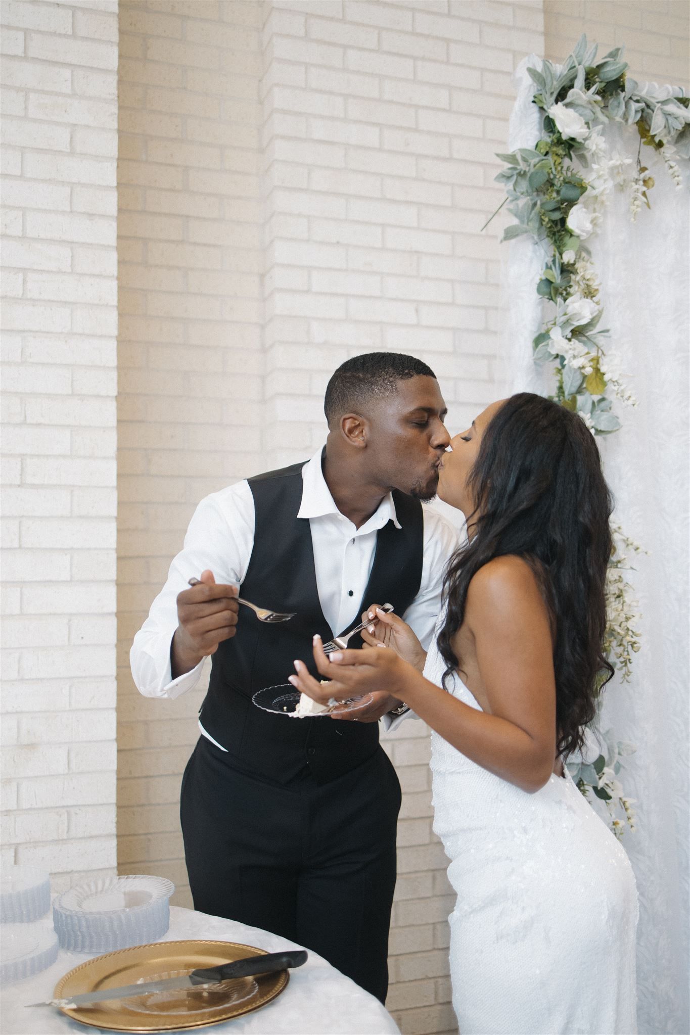 Newly married couple shares a kiss while eating wedding cake together in front of white brick wall with floral decor.