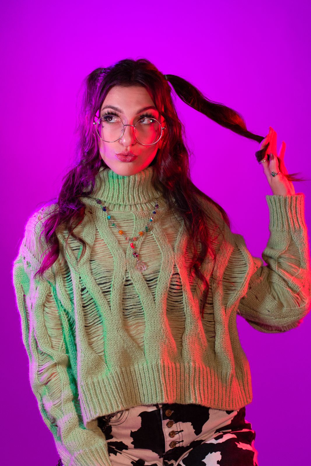 Portrait photography: a woman wearing large glasses and a green sweater twirls her hair surrounded by purple and red light