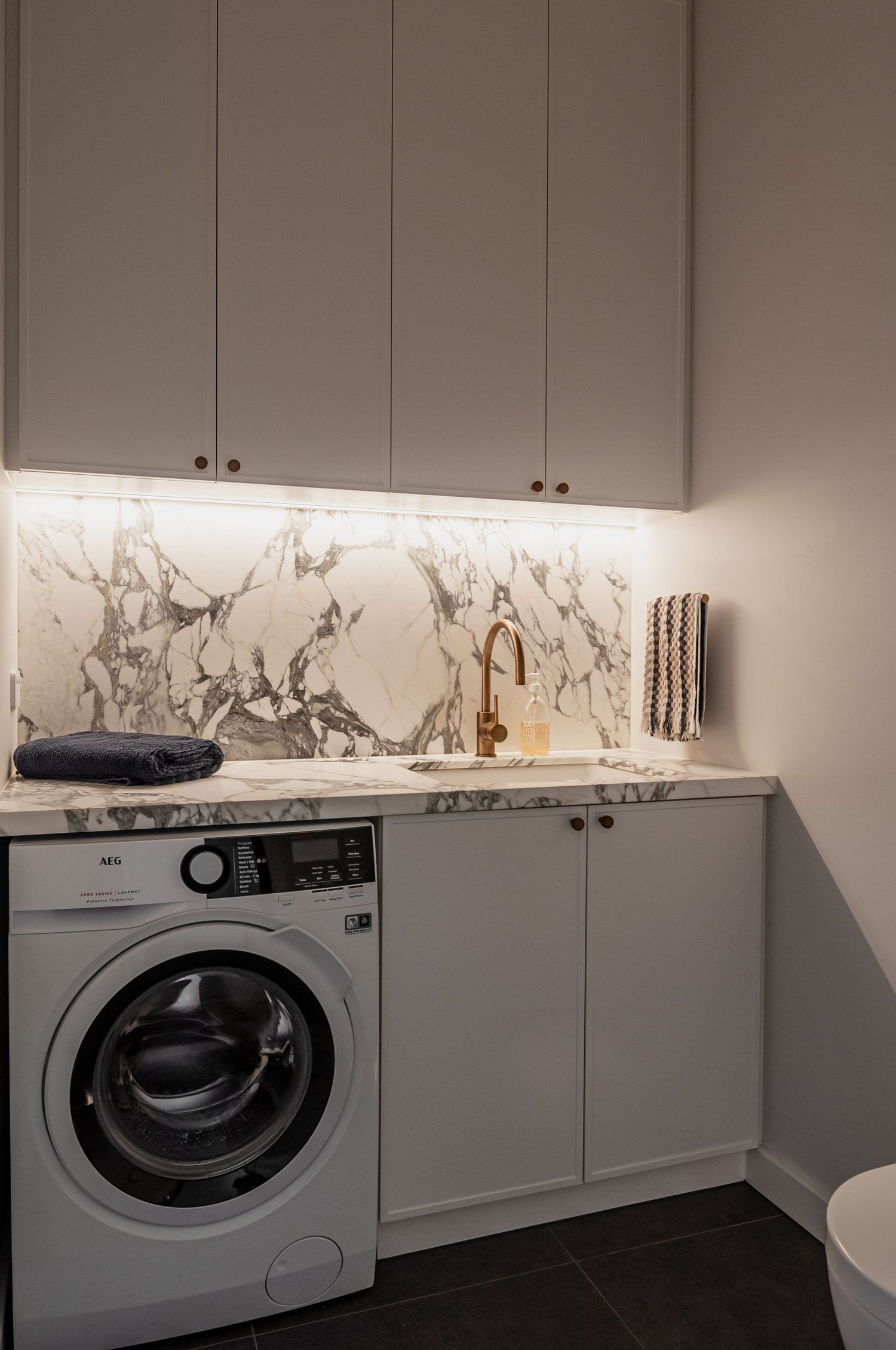 Modern laundry room with grey cabinets, marble backsplash, and front-loading washing machine.