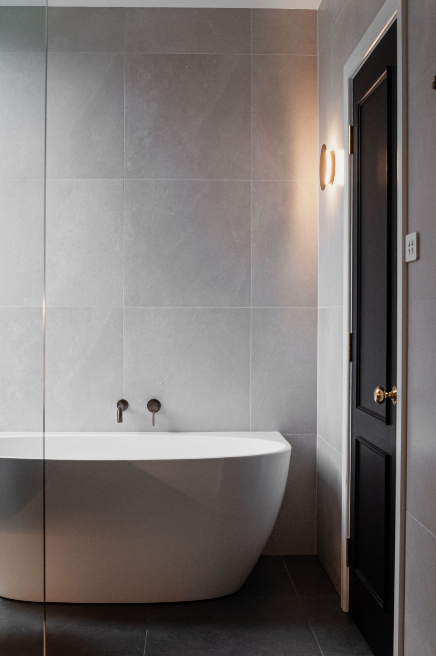 Minimalist bathroom featuring freestanding tub, grey tile walls, and black door.